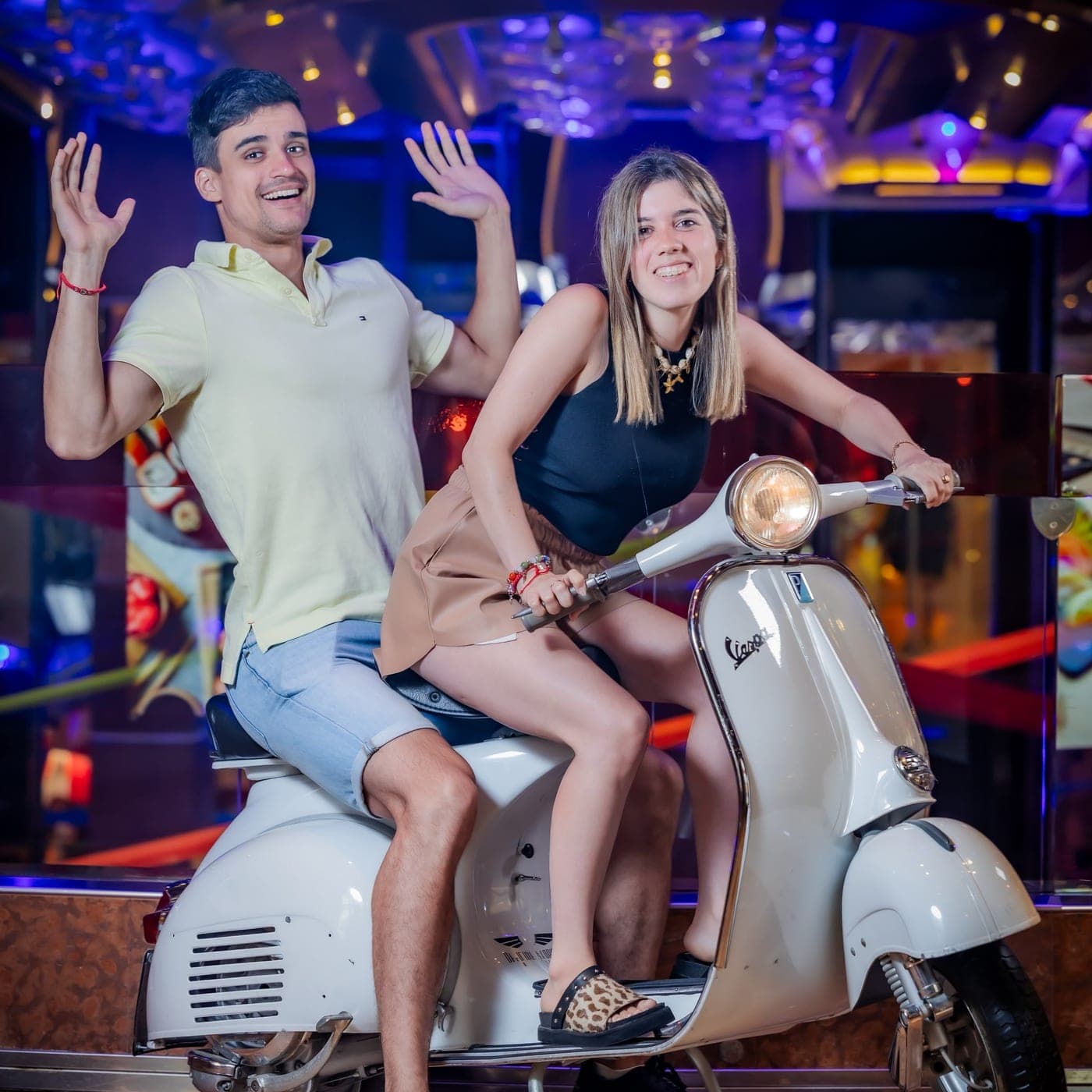 Caro y Valen saludando arriba de una Vespa blanca, entre luces de colores